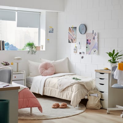 Bedroom with white brick walls, beige bedding set, soft pillow, comforter, under window, desk, chair, plants.