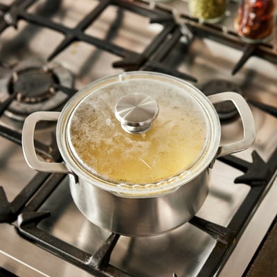 A saucepan on a stove with boiling liquid and a glass lid, featuring a built-in strainer.