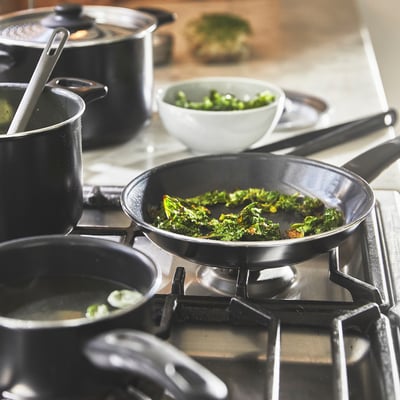 On a stovetop, various pots and pans cook greens. One pan details even heat distribution due to thick bases, ideal for cooking.
