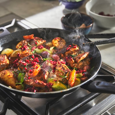 Large frying pan cooking chicken, veggies, and pomegranate seeds on a stove, steam visible, handles on either side.