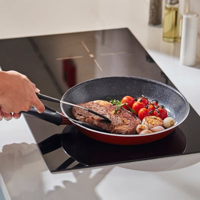 Hand holds tongs flipping steak in red non-stick pan on induction hob, surrounded by veggies.