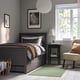 Dark grey HEMNES bed, white/beige bedding. Black bedside table, lamp, green view. Beige chair, orange pillow, striped rug.