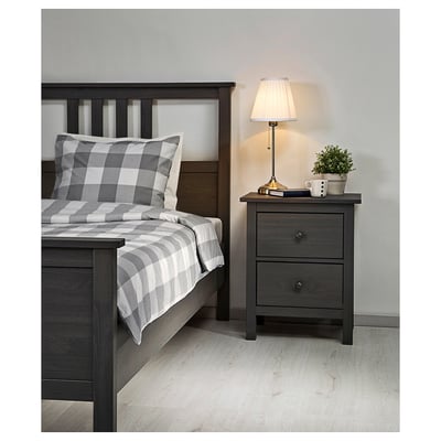 Bedroom scene: dark grey wooden bed with grey chequered bedding, bedside table with lamp, plant, and mug.