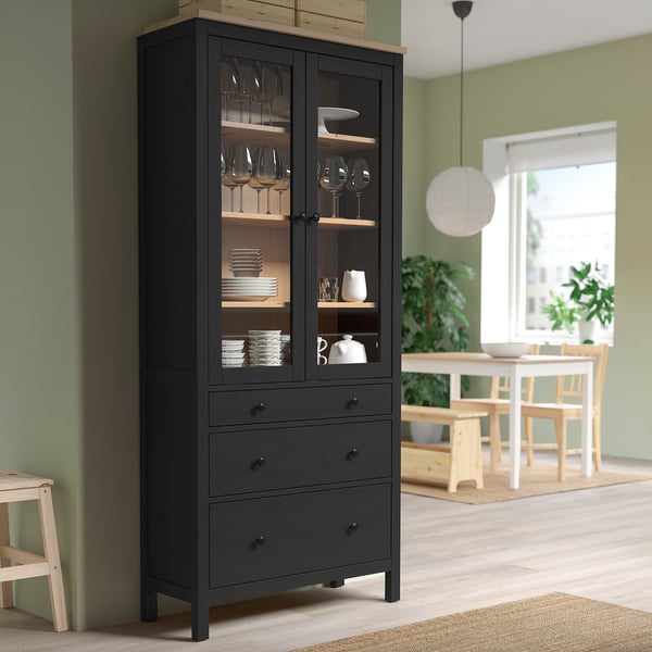 Black HEMNES cabinet with glass doors, wooden shelves, and glass/dishware. Tall near green wall.