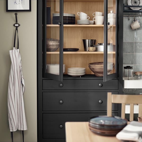 Black HEMNES cabinet with glass doors, holding plates, bowls, and mugs, striped apron, kitchenware.