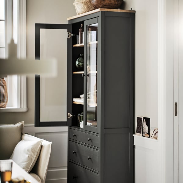 A dark HEMNES cabinet with glass doors, standing tall beside a door. Its made of solid wood, tall with adjustable shelves, neatly storing glassware.