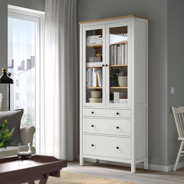 Wooden HEMNES cabinet with glass doors, storing books and items, between window and chair.