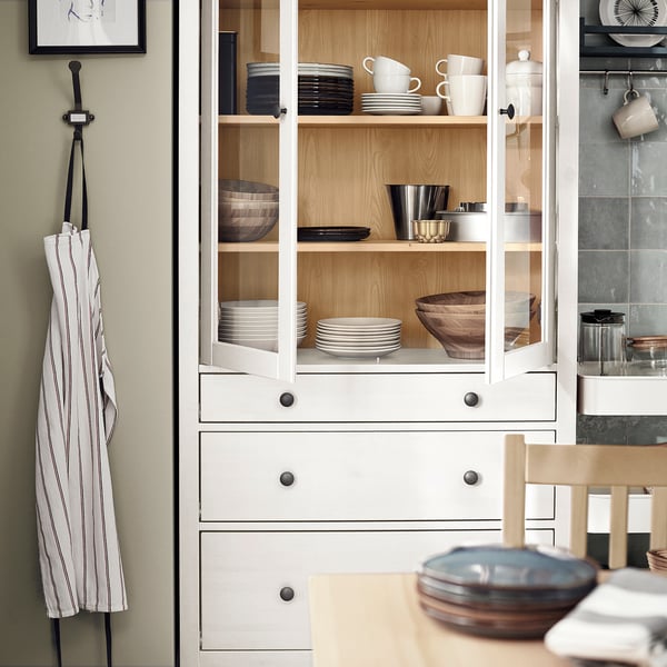 HEMNES white wooden cabinet, with glass doors, holds stacked dishes, wooden bowls, and cups. An apron hangs beside it, emphasising its size and style.