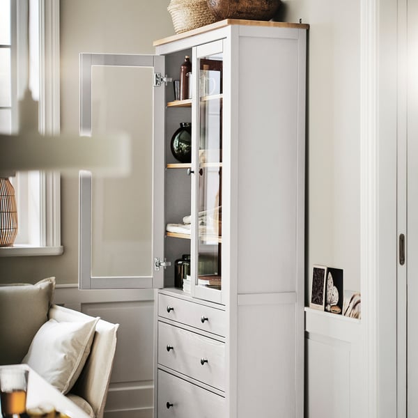 A tall HEMNES wooden cabinet, white, with glass doors and drawers. It displays decorative items inside and stands near a staircase and chair.
