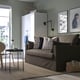 Sofa bed in grey fabric with patterned cushions sits in a modern room with a tall white cabinet behind. Next to a small table holding vases.