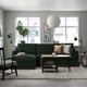 Modern living room: large green l-shaped sofa, coffee table, large rug, potted plant, framed art, black floor lamp, hanging light, chair with blanket.