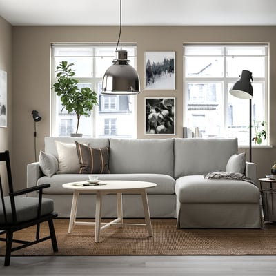 A modern living room with a grey sectional sofa, white coffee table, and black floor lamp. Large windows, two artworks, plant, rug, and wooden chair.