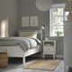 Minimalist bedroom with white bed, grey linens, yellow pillow. Art, bedside table, lamp, books, plant, rug, basket. Cosy and organised.