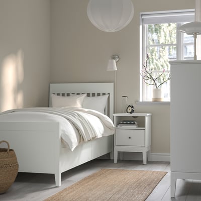 Modern minimalist bedroom with IDANÄS white bed, bedside table, and rug. Soft lighting, plants by window.
