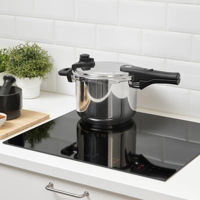 A sleek black pressure cooker with two handles sits on an induction hob, surrounded by white kitchen decor.