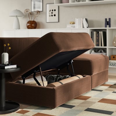 Brown modular sofa with ottoman, open storage for blankets and items, in a cosy living room setting with decor and shelves.