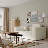 Modular sofa in white l-shape, with cushions and chaise. On earthy rug, near coffee table and lamp. Minimalist room with art and cabinet.