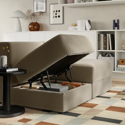 Beige modular sofa with storage compartment open, revealing blankets and books inside. Shelves with decor in background.
