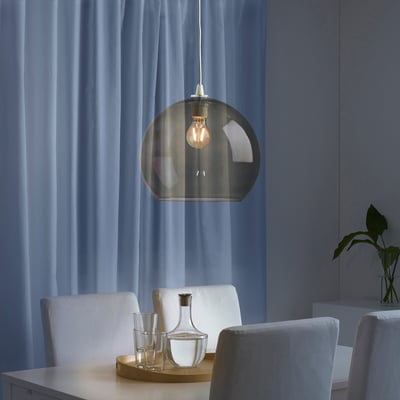 Modern dining setup with smoked glass light, white chairs, clear glasses, wooden tray, and vase. Curtains drawn.