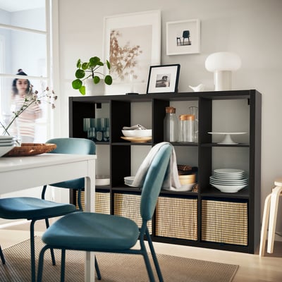 Cosy dining room with blue chairs, black KALLAX storage unit, white dishes, glassware, plants, and wall art.