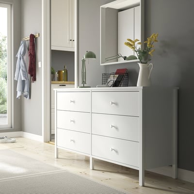 White 6-drawer KILBERGET chest under mirror, left with clothes, top with lamp and yellow flowers, traditional design.