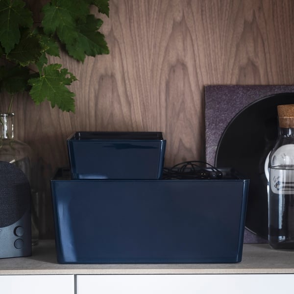 Two blue storage boxes on a shelf, one with a lid, next to a speaker and a bottle.