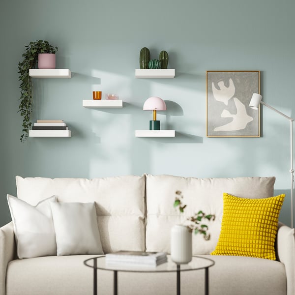 Modern living room: blue walls, plants, books, pink lamp, white couch, yellow pillow, round table, abstract art.
