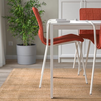 Modern office setting with white desk and orange chairs, one pushed in. A large potted plant and a beige rug are also visible.