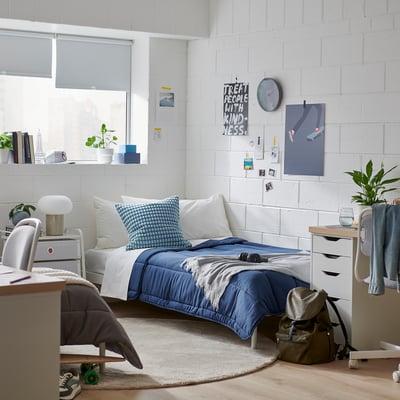 Cosy bedroom with a LÅSBRÄKEN blue comforter on a single bed. Plants, books, and a backpack surround, creating a lived-in yet organised space.