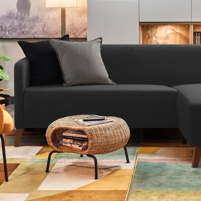 Modern living room with a black l-shaped sofa, grey pillows, a wicker ottoman with books, orange chair, colourful rug, and lit display cabinet.