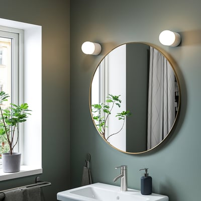 Modern bathroom with large gold-framed mirror above white sink, reflecting plant and curtain, lit by two circular wall lights, green walls.