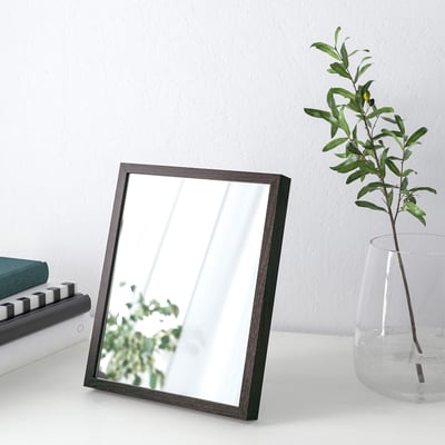 Minimalist mirror on table, reflecting window, flanked by books and vase with greenery.