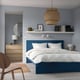 Bedroom with blue MALM bed, white linens, wooden dresser, mirror, pendant lamp, bookshelf with books, wooden chair, and floor lamp.