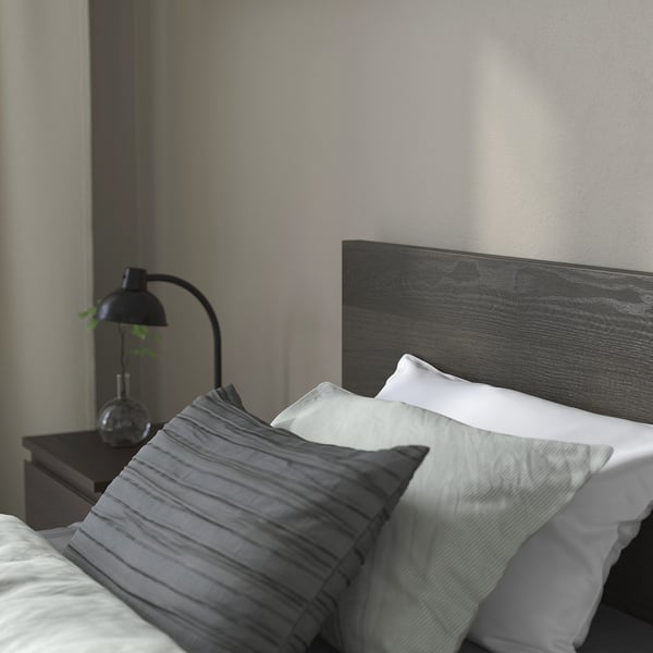 A minimalist bed with a dark wooden headboard, white and grey pillows, and a black lamp with a plant on a bedside table.