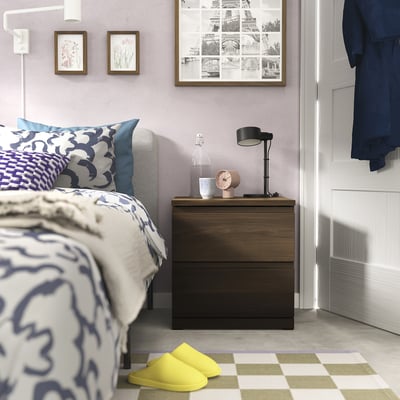 Bedroom with modern bed in dark wood, blue patterned sheets, yellow slippers, and a black bedside table with a lamp, clock, and framed wall art.