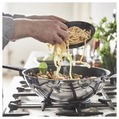 A hand adding noodles from a bowl to a silver wok on a gas stove with flames. Green veggies and a plant are in the background.