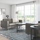 Modern grey sofa with white pillows, round glass table, large windows, stylish decor.