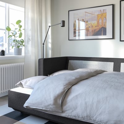 Modern bedroom with grey sofa bed, bridge artwork, tall plant, and sheer curtains.