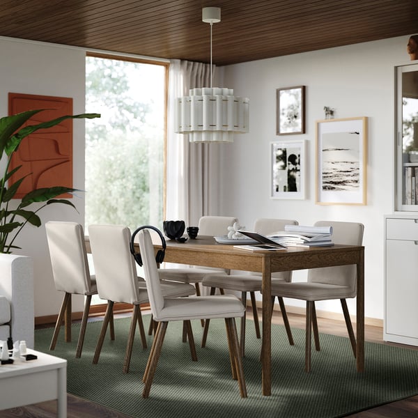 Modern dining room with MORUM rug, wooden table, beige chairs, flowers, chandelier, artwork, clock, window, plant, cabinet.