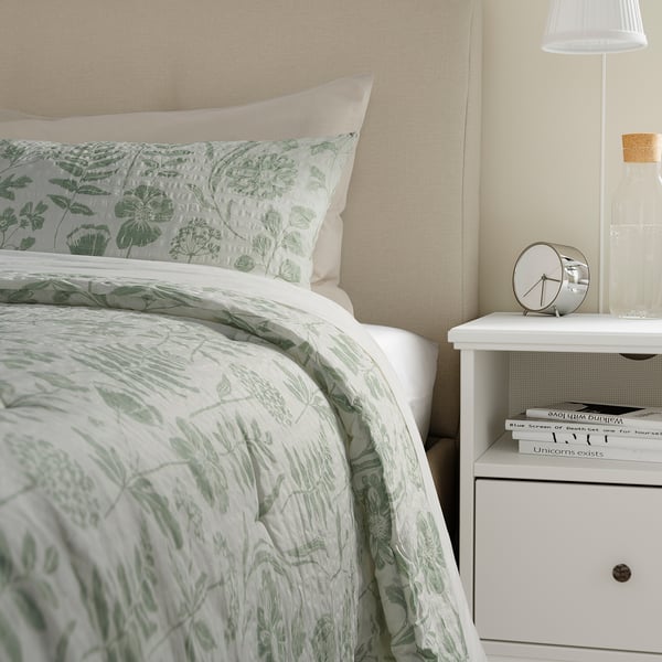 Bed with NÅLBJÖRNBÄR sage green floral comforter and pillow sham, near bedside table with books and clock.