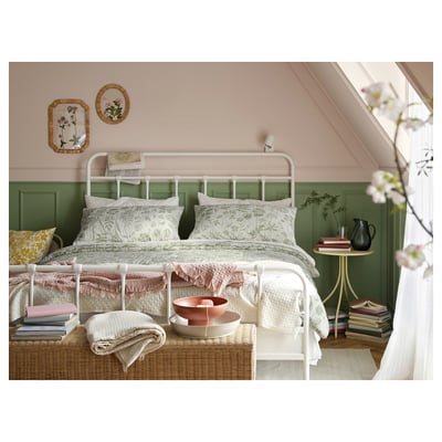 Cosy bedroom with white bed, floral bedding, green wall panels, rattan chest, books, and pink decor.