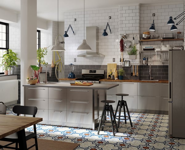Modern kitchen with stainless steel NOGSTA fronts, patterned floor tiles, hanging lights, and black stools.