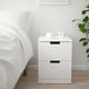 A white ikea NORDLI nightstand with two drawers next to a bed, holding a coffee cup and digital clock. Minimalist scandinavian design.