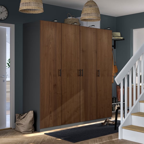 Large wooden brown PAX wardrobe near white staircase. Two woven lampshades hang above. A black mat and glimpse of outdoor space beside white door.