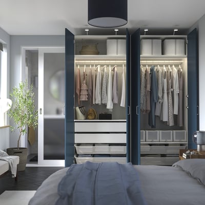 Modern bedroom with open wardrobe showcasing organised clothing, shelves, and drawers in a PAX system. Integrated lighting highlights stored items.