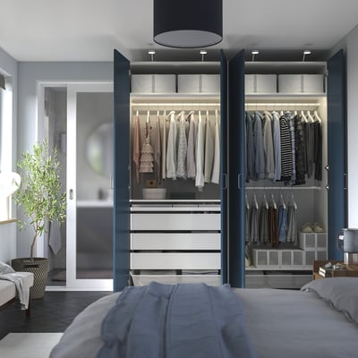 Modern bedroom with open PAX wardrobe showcasing clothes, drawers, and storage boxes. Soft lighting, grey walls, and a blue bedspread.