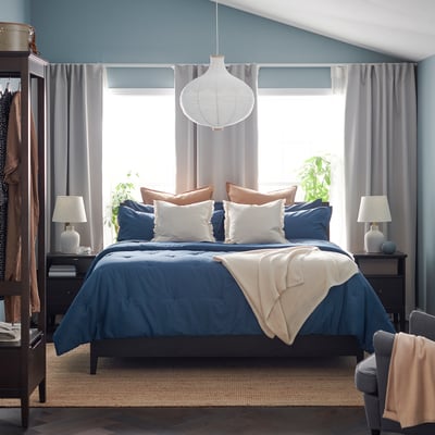 Bedroom with blue PIGGTISTEL comforter on bed, two nightstands, lamps, large window with curtains, hanging coat rack, and chair.