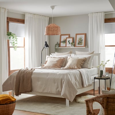 Cosy bedroom with large bed, beige linens, natural light, wicker light, art, flowers, and blanket.