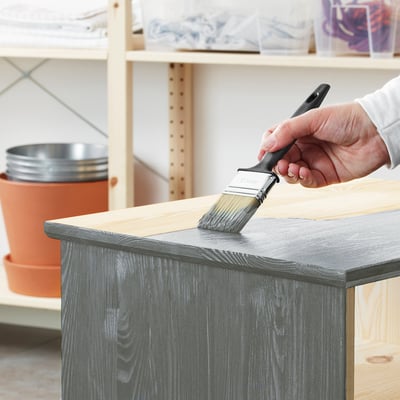 Person applying grey paint to wooden furniture using brush. Paint is water-borne, low gloss, suitable for indoor use.
