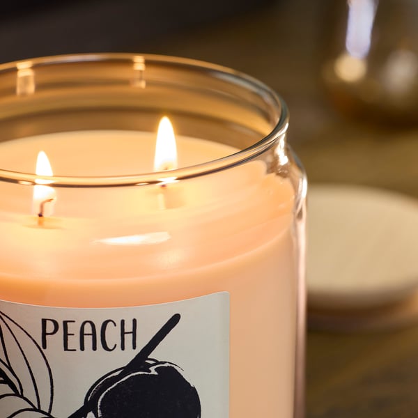 Peach-scented candle with 3 wicks glowing on wooden table.
